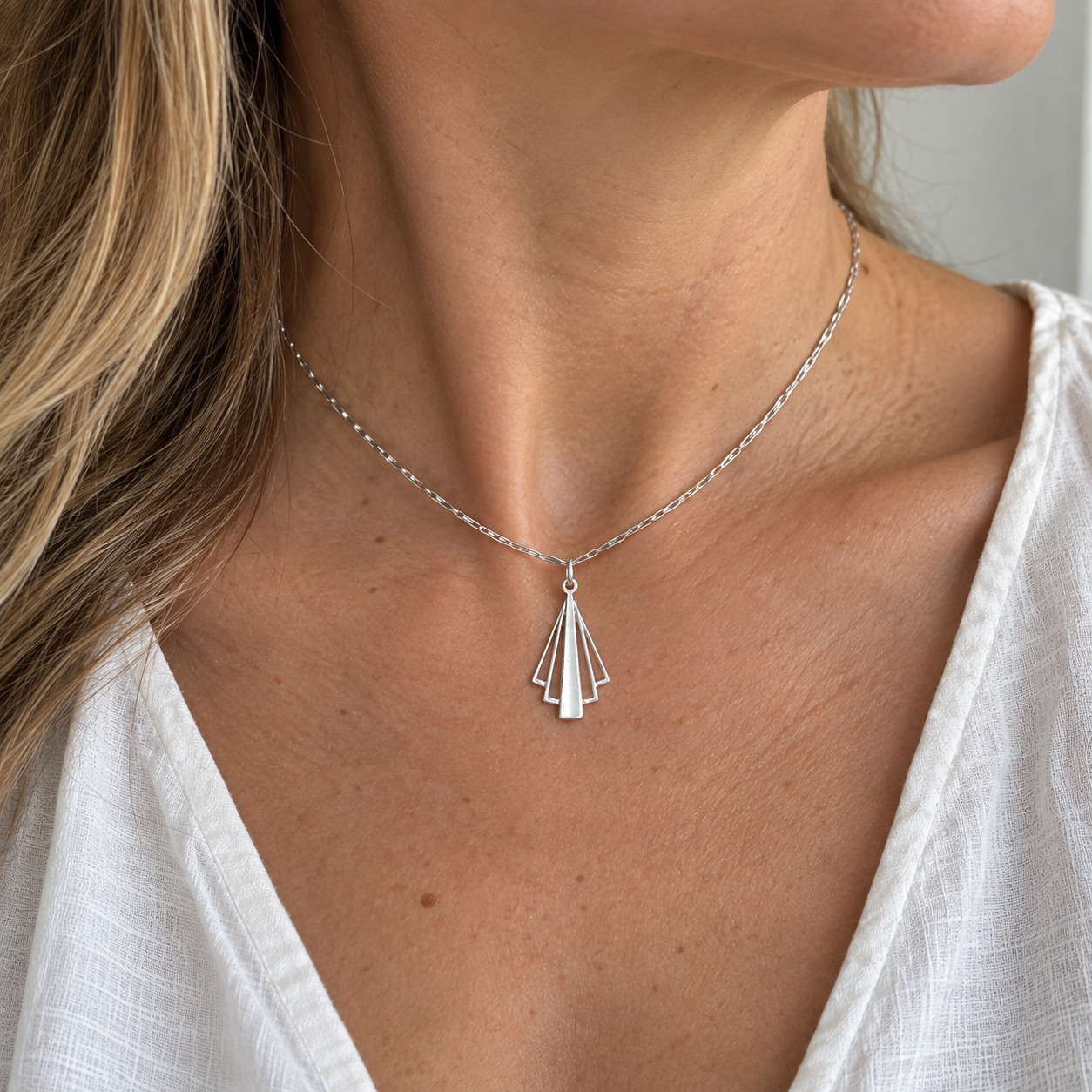 A close-up of a woman wearing a delicate silver chain necklace with a geometric fan-shaped pendant, paired with a white top.