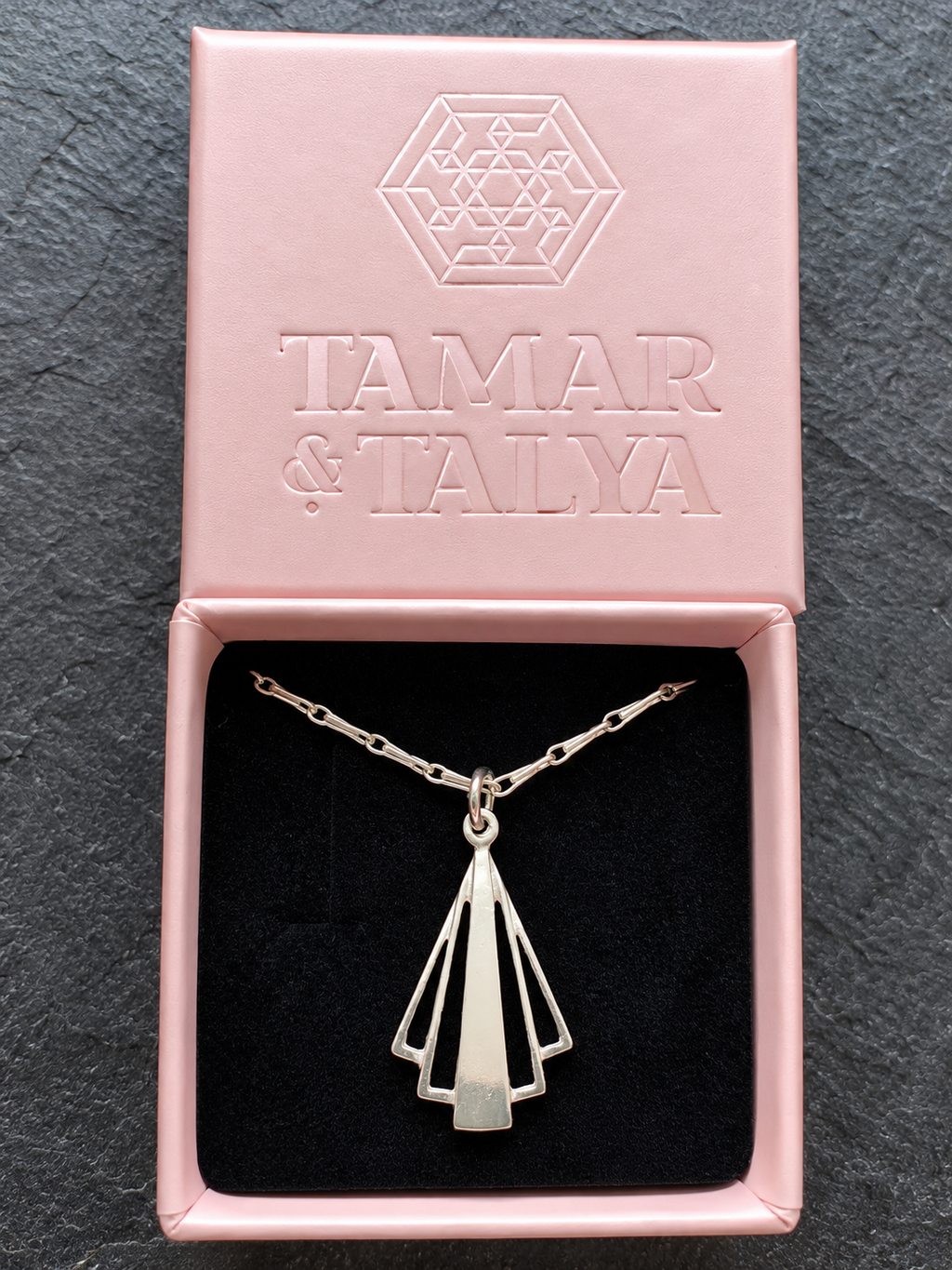 A silver geometric pendant necklace displayed inside an open pink jewelry box with a black velvet insert. The embossed lid reads 'TAMAR & TALYA'.