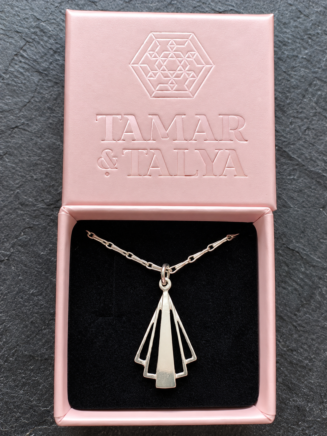 A silver geometric pendant necklace displayed inside an open pink jewelry box with a black velvet insert. The embossed lid reads 'TAMAR & TALYA'.