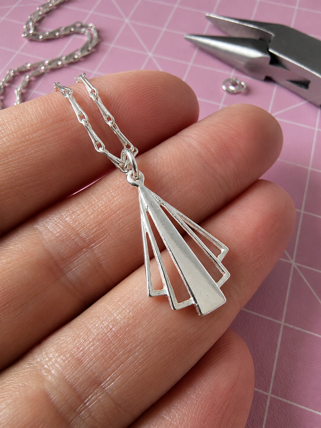 A silver geometric pendant necklace held in a person's hand, featuring a fan-like layered design on a chain, with pink cutting mat and metal pliers in the background.
