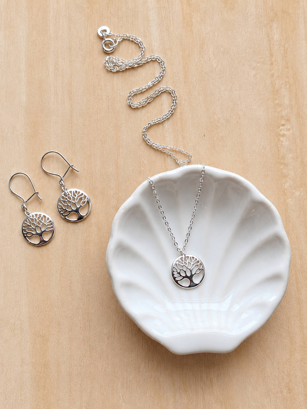Silver tree of life jewelry set including a necklace and matching drop earrings, displayed on a white scalloped dish over a light wood background.
